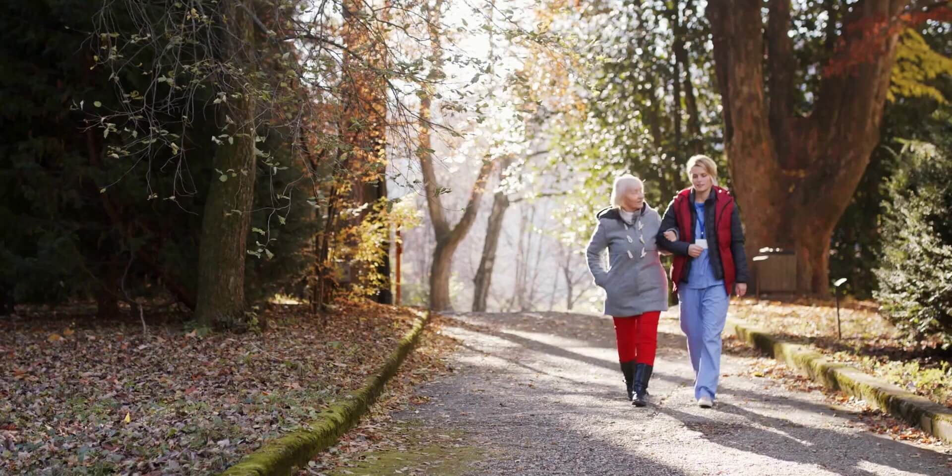 caregiver and a senior walking arm in arm down an autumn lane