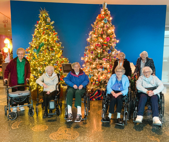 Elderly women in wheelchairs pose in front of illuminated Christmas trees.